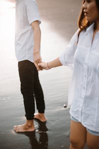 A couple holding hands and walking barefoot on a serene beach at sunset.